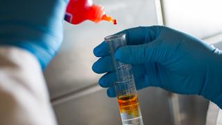 A pair of gloved hands in a chemical expermenting area is adding an orange liquid from a squeeze bottle into a flask holding a clear substance.